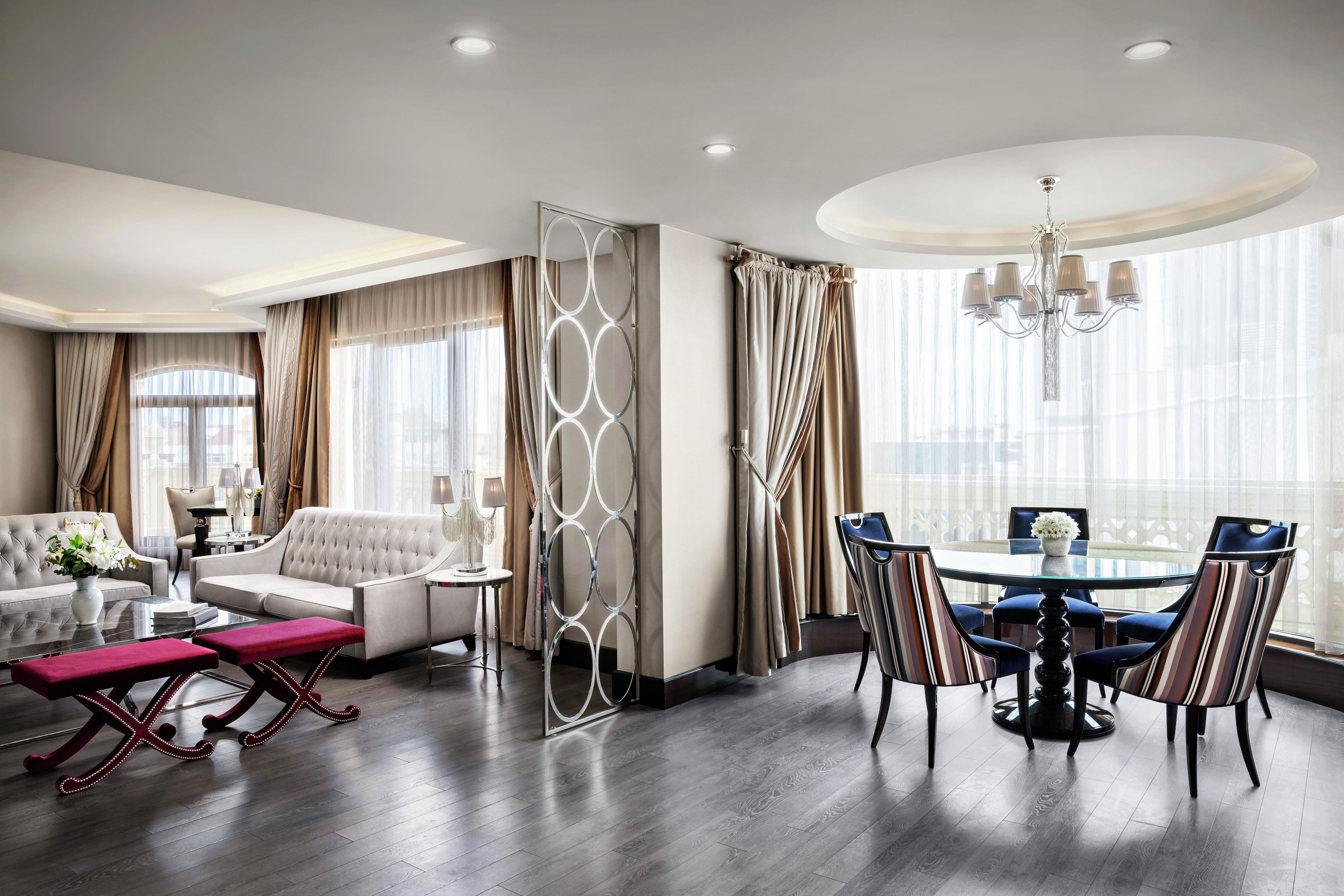 A modern hotel room with a dining area, a sofa, a coffee table, and a view of the city.