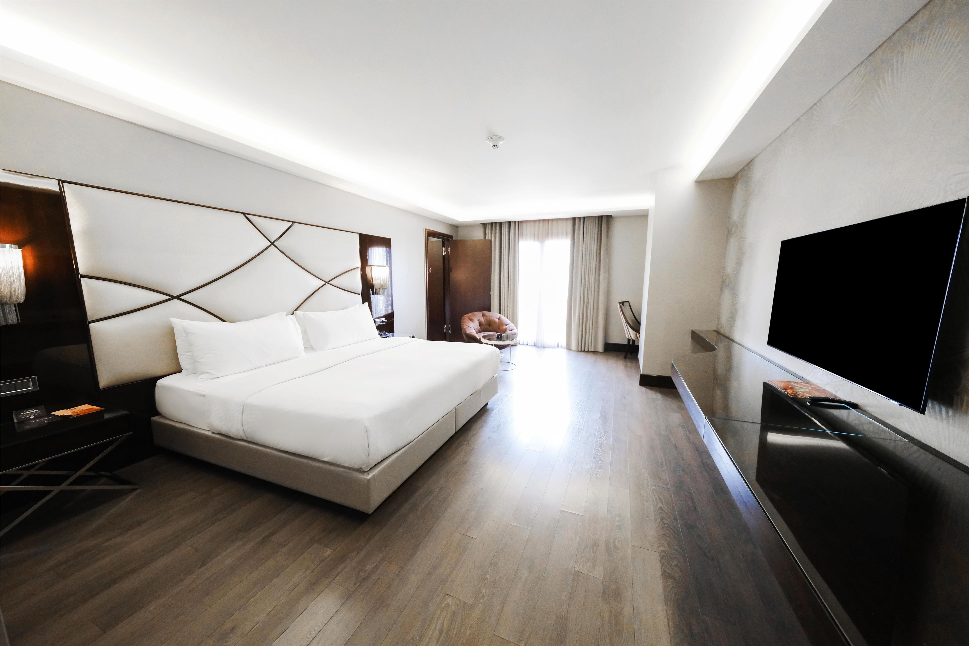 A modern hotel room with a large bed, a flat-screen TV, a desk with a lamp, and a chair.