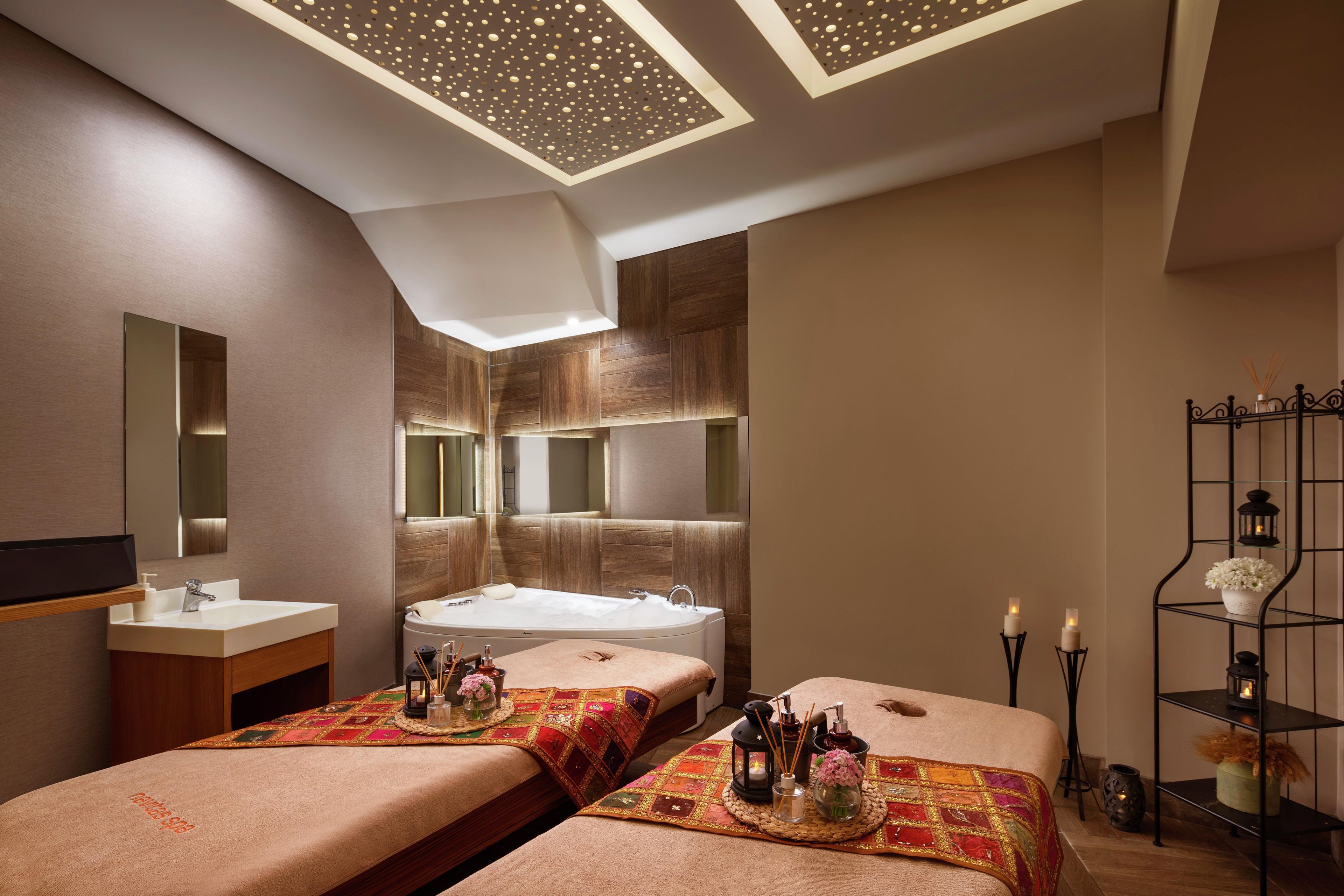 A modern spa room with a large bathtub, two massage tables, a vanity with a mirror, and a ceiling with a geometric pattern.