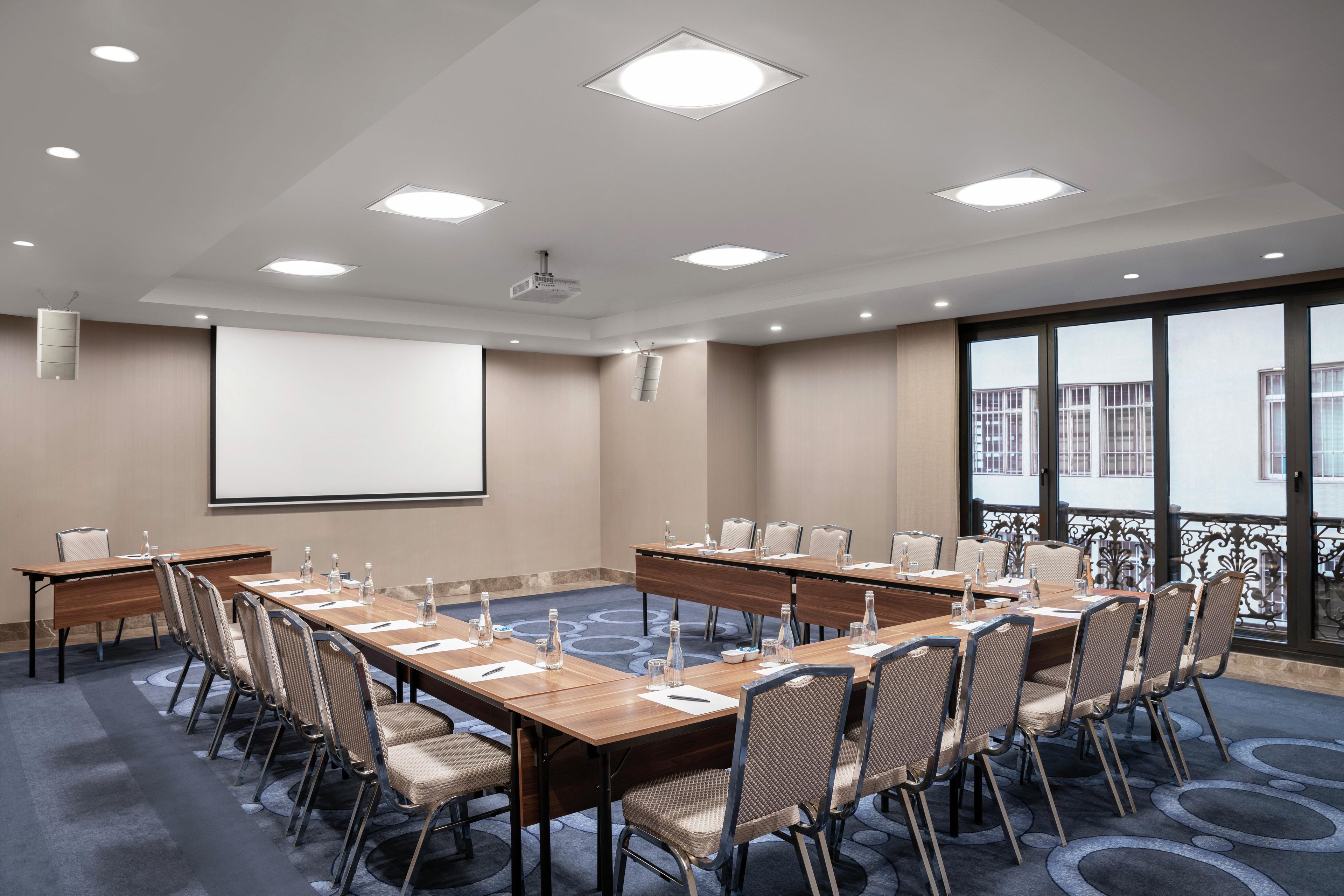 Une salle de conférence équipée d’une grande table, de chaises, d’un écran de projection et d’une grande fenêtre.