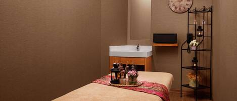 A spa room with a bed, a sink, a mirror, and a shelf with candles and flowers.