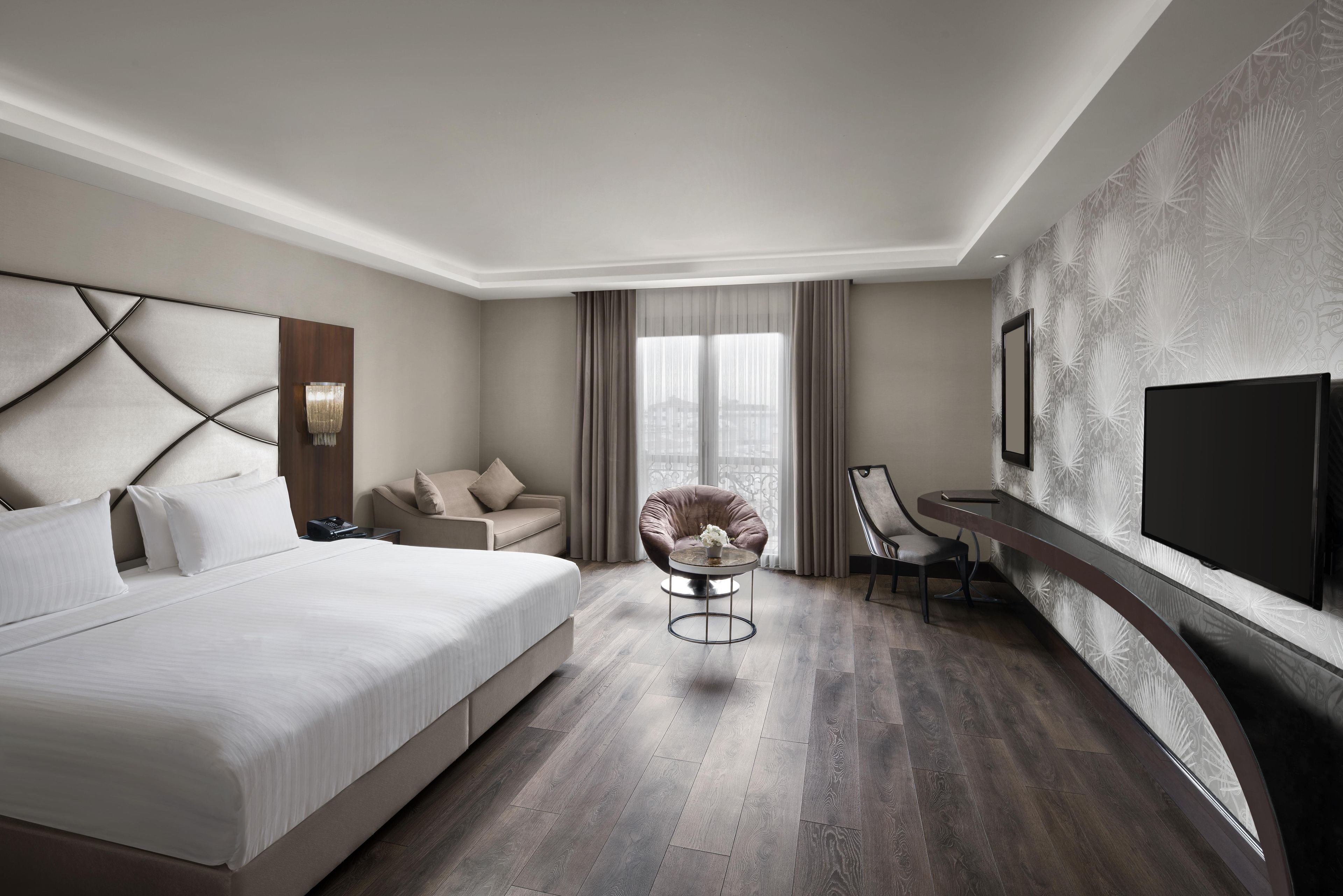 A modern hotel room with a large bed, a seating area with a chair, a small table, and a TV mounted on the wall.