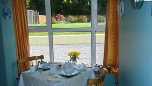 A dining table set for two with a view of a garden through large windows. - Ashfield Bed & Breakfast (Kenmare)