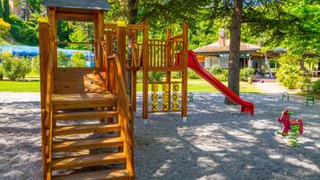 A playground with a wooden slide and climbing structure, surrounded by trees and a building in the background.