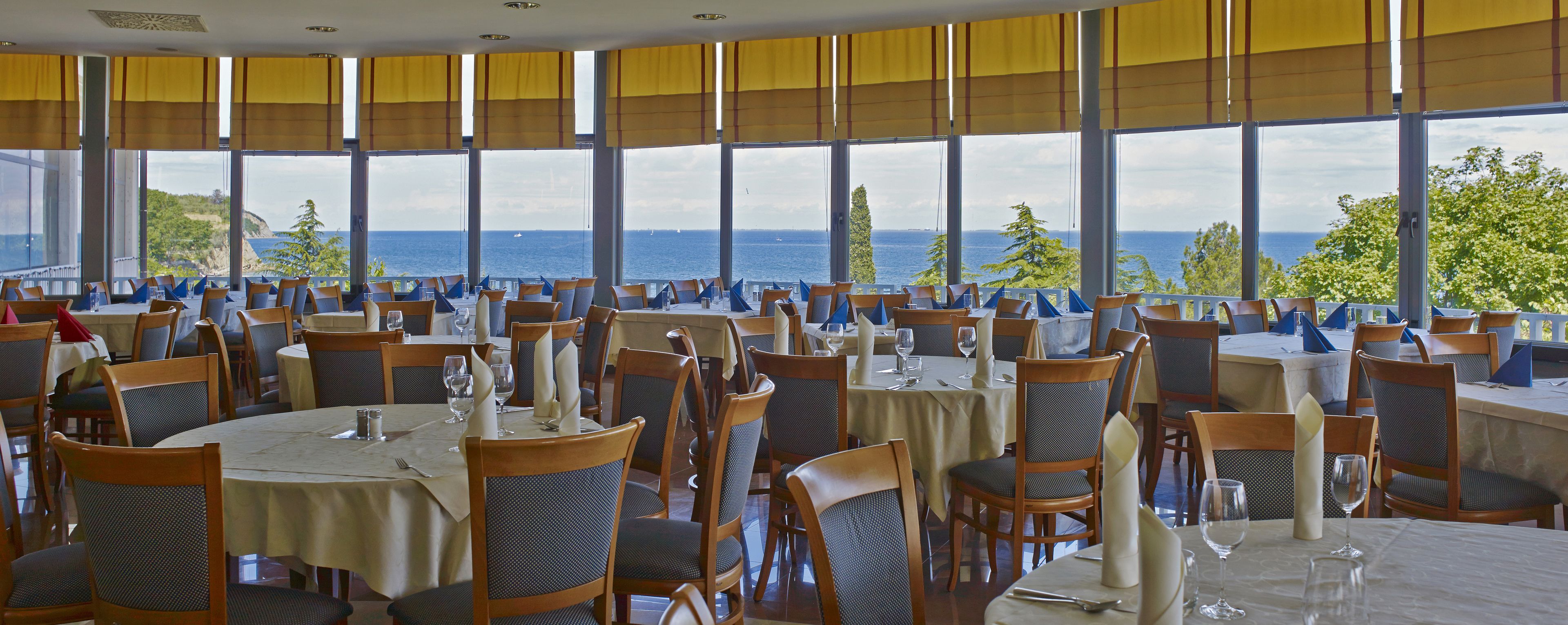 Un ristorante con tavoli e sedie rotonde, ampie finestre vista mare e tende gialle.