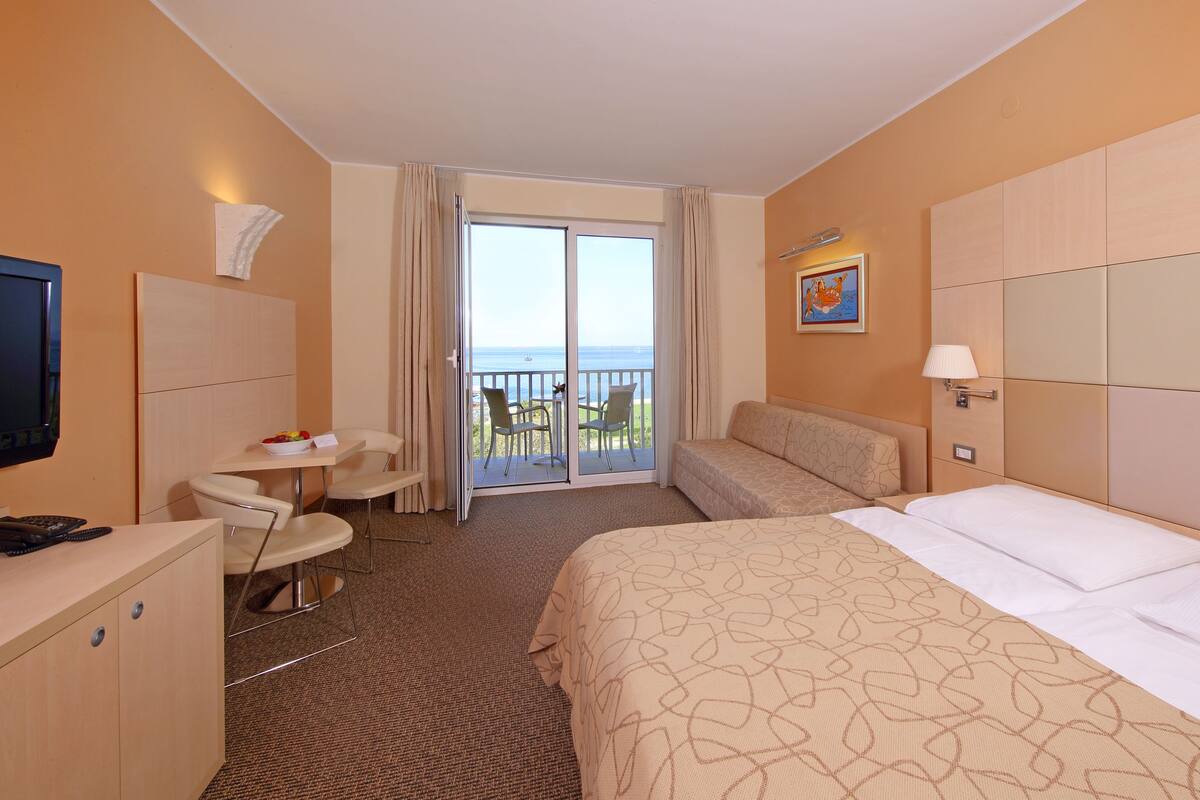 Une chambre d’hôtel comprenant un lit, un bureau, une chaise, une télévision, une fenêtre donnant sur la mer et un tableau accroché au mur.