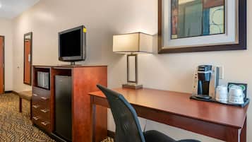 A hotel room with a wooden desk, a television, a lamp, a coffee maker, and a refrigerator.