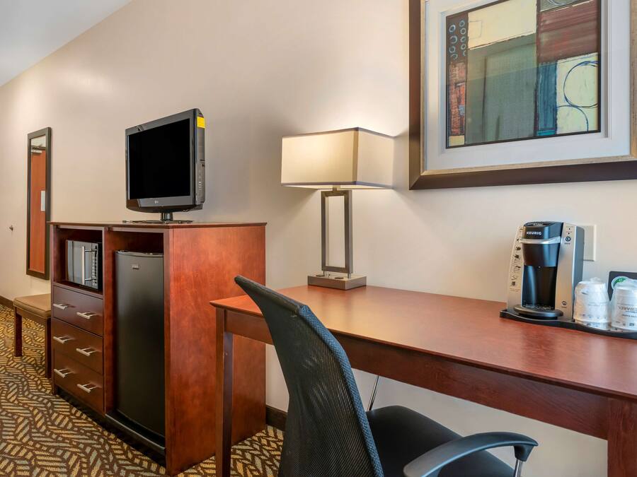 A hotel room with a wooden desk, a television, a lamp, a coffee maker, and a refrigerator.