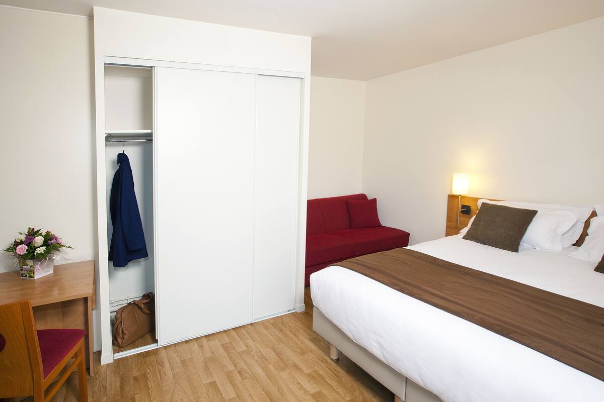 Een hotelkamer met een bed, een rode bank, een houten bureau met stoel, en een witte inloopkast met jassen.