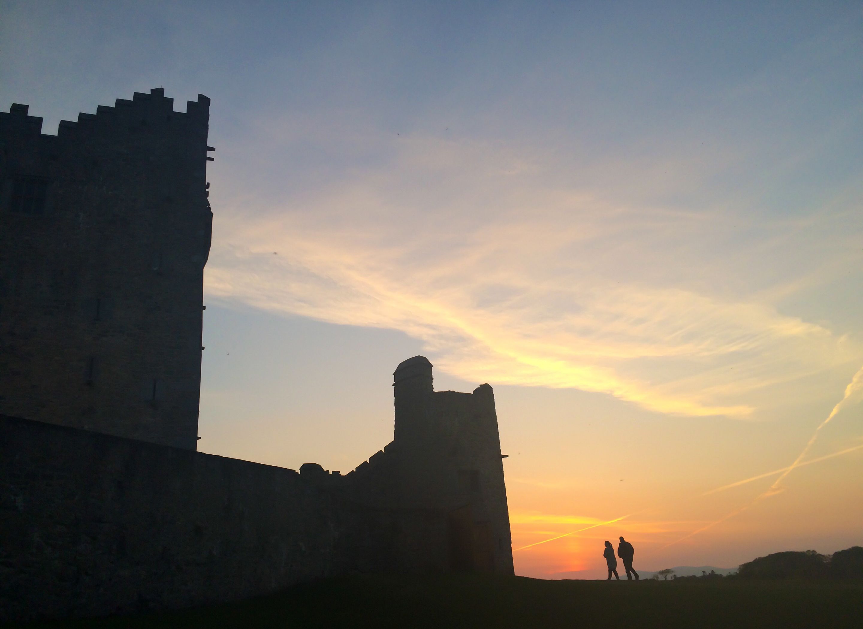 A historical stone structure with a prominent tower and a smaller building, set against a sunset sky.