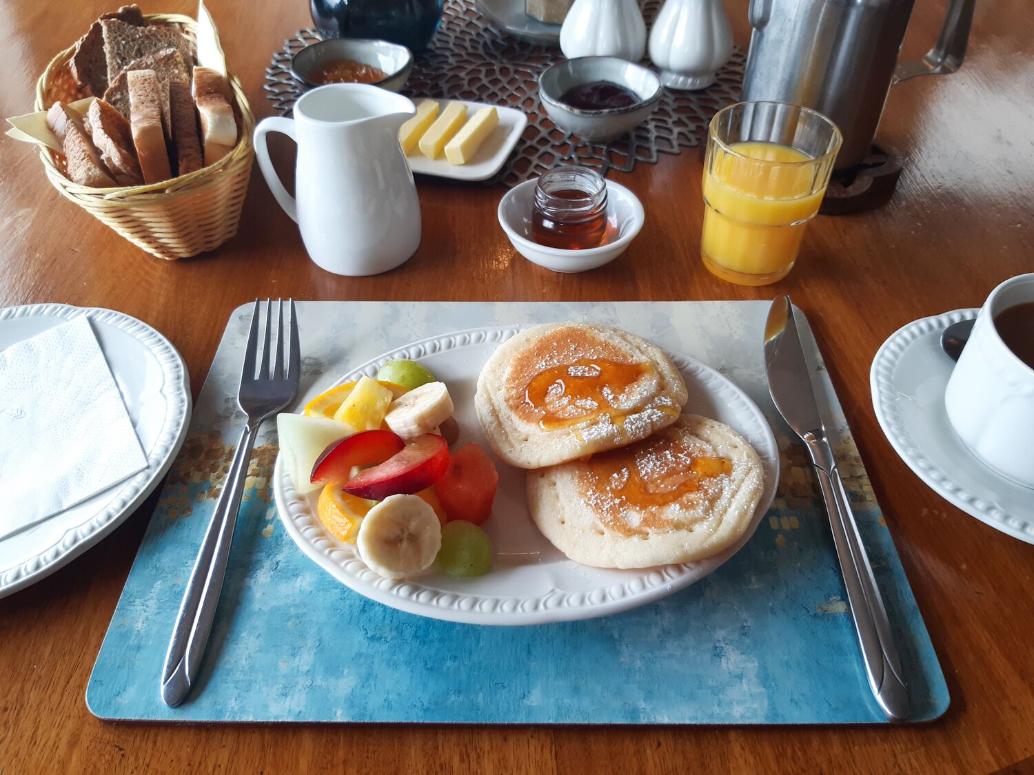A breakfast setting with pancakes, fruit, butter, and a coffee cup on a wooden table.