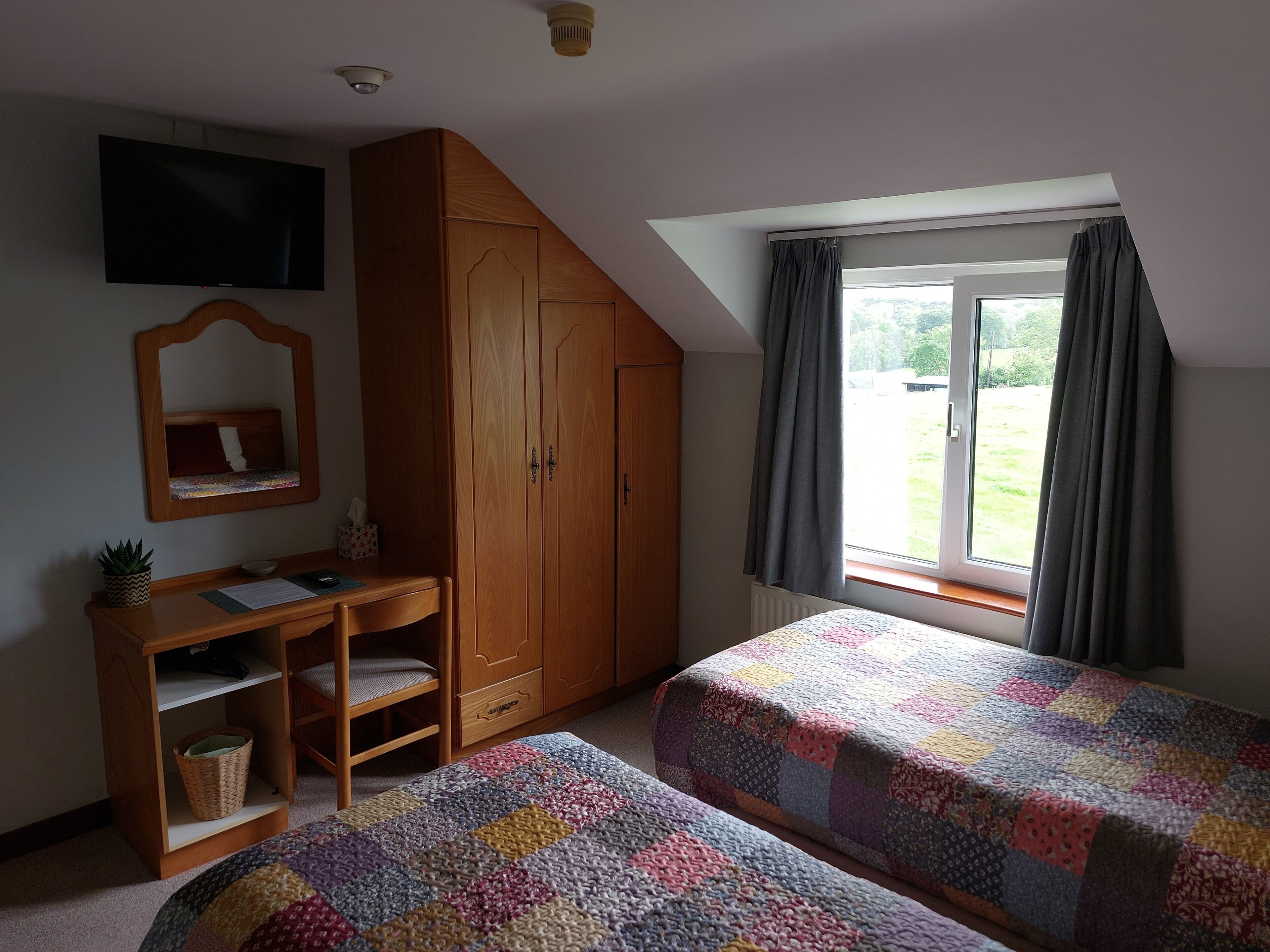 A bedroom with two beds, a desk, a wardrobe, a mirror, and a window with curtains.