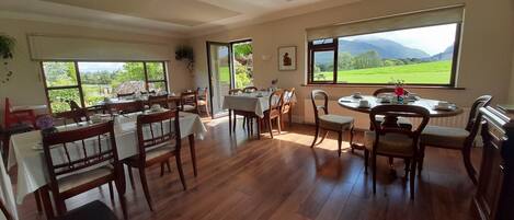 Un espace repas avec des tables et des chaises en bois, de grandes fenêtres offrant une vue sur un paysage verdoyant et des montagnes.