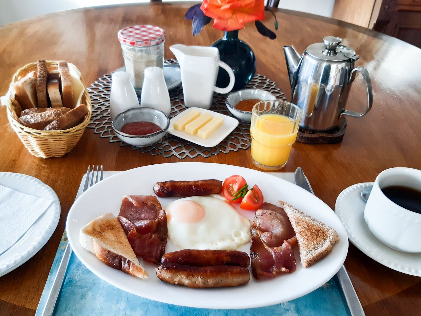Ein Frühstückstisch mit Eiern, Würstchen und Toast, sowie Beilagen, einer Kaffeetasse und einem Glas Fruchtsaft.