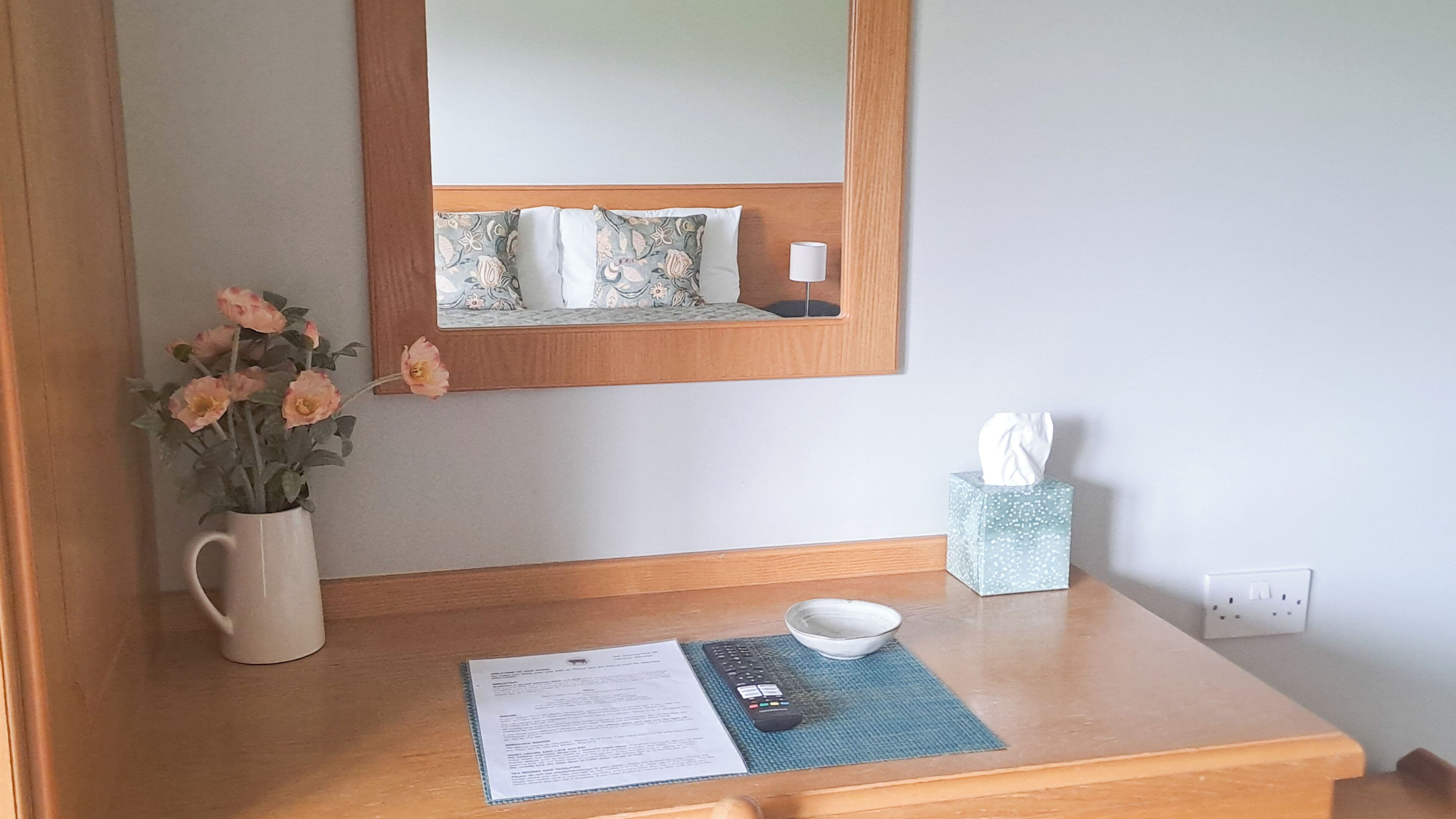 Une table de chevet en bois, avec une télécommande, une boîte de mouchoirs, un vase avec des fleurs et un miroir au-dessus de la table.