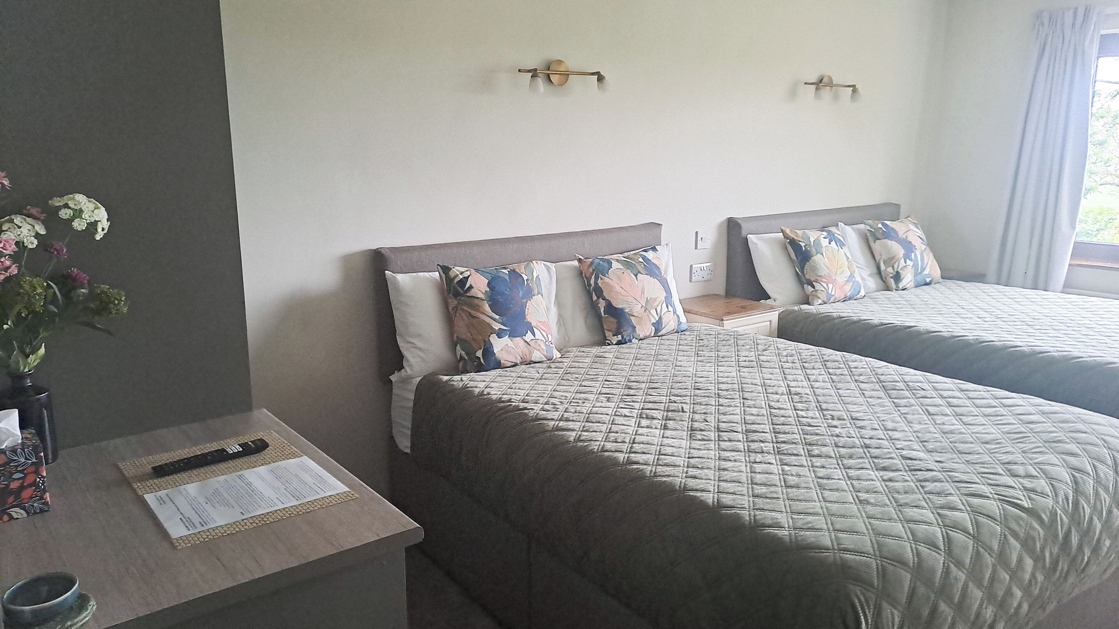 A hotel room with two beds, a nightstand, a vase with flowers, and a window with curtains.