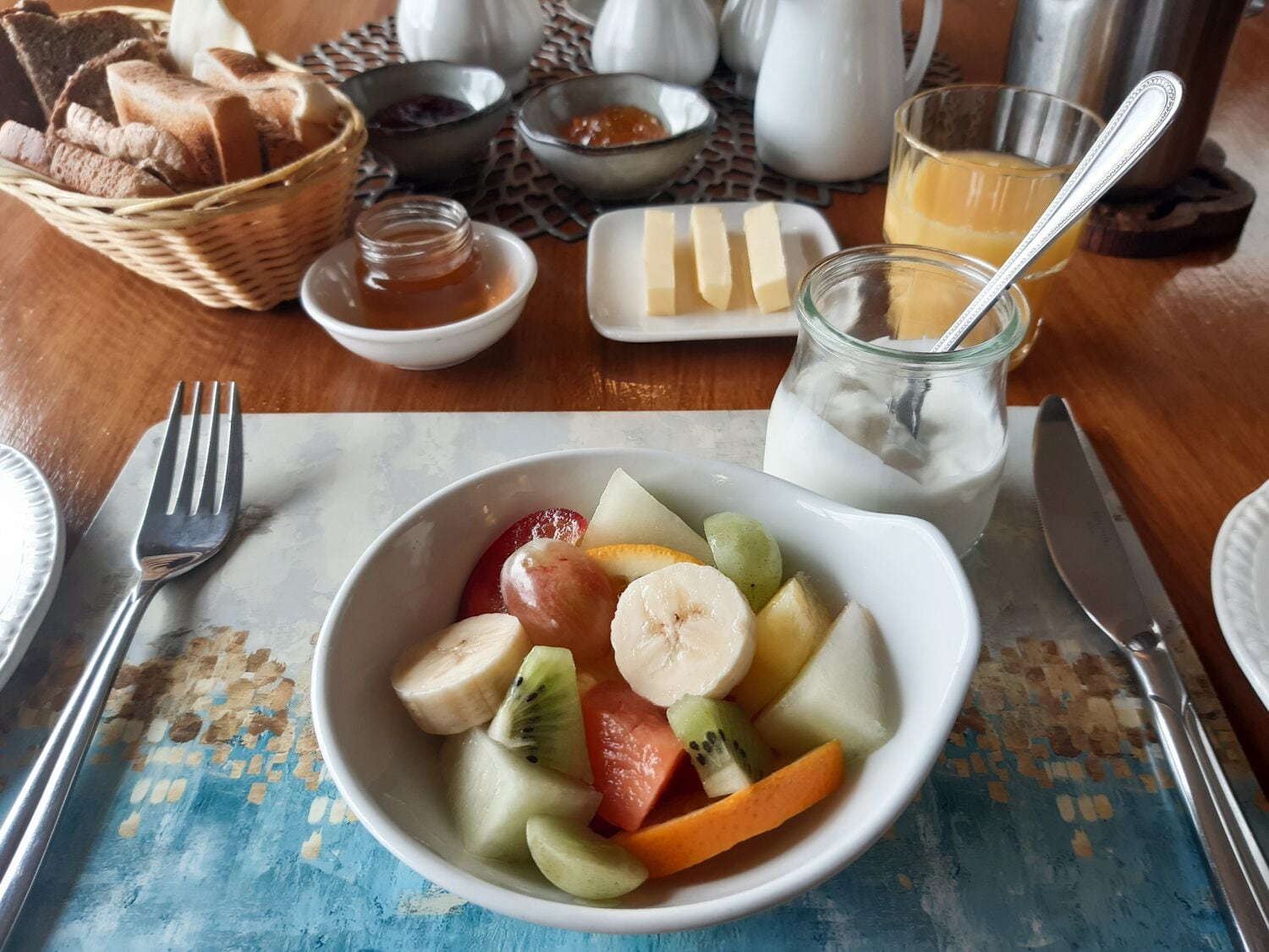 Un desayuno con un tazón de fruta, pan, mantequilla, mermelada y un vaso de jugo de naranja.