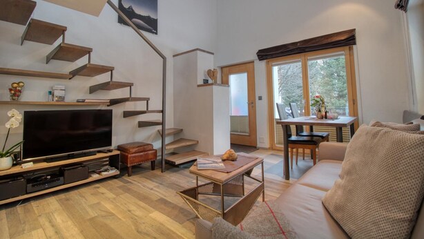 A modern living room with a flat-screen TV, wooden flooring, a sofa, a coffee table, a staircase, and a dining area with a table and chairs. - Chalet Abacus (Zermatt)