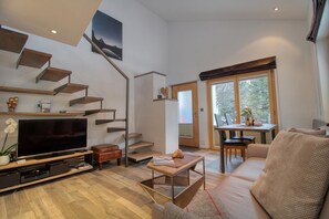 Una sala de estar moderna con una televisión de pantalla plana, piso de madera, un sofá, una mesa de centro, una escalera y un comedor con una mesa y sillas.