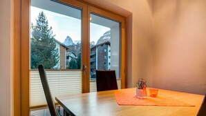 A room with a wooden table, a chair, and a window with a view of buildings and trees. - Chalet Abacus (Zermatt)