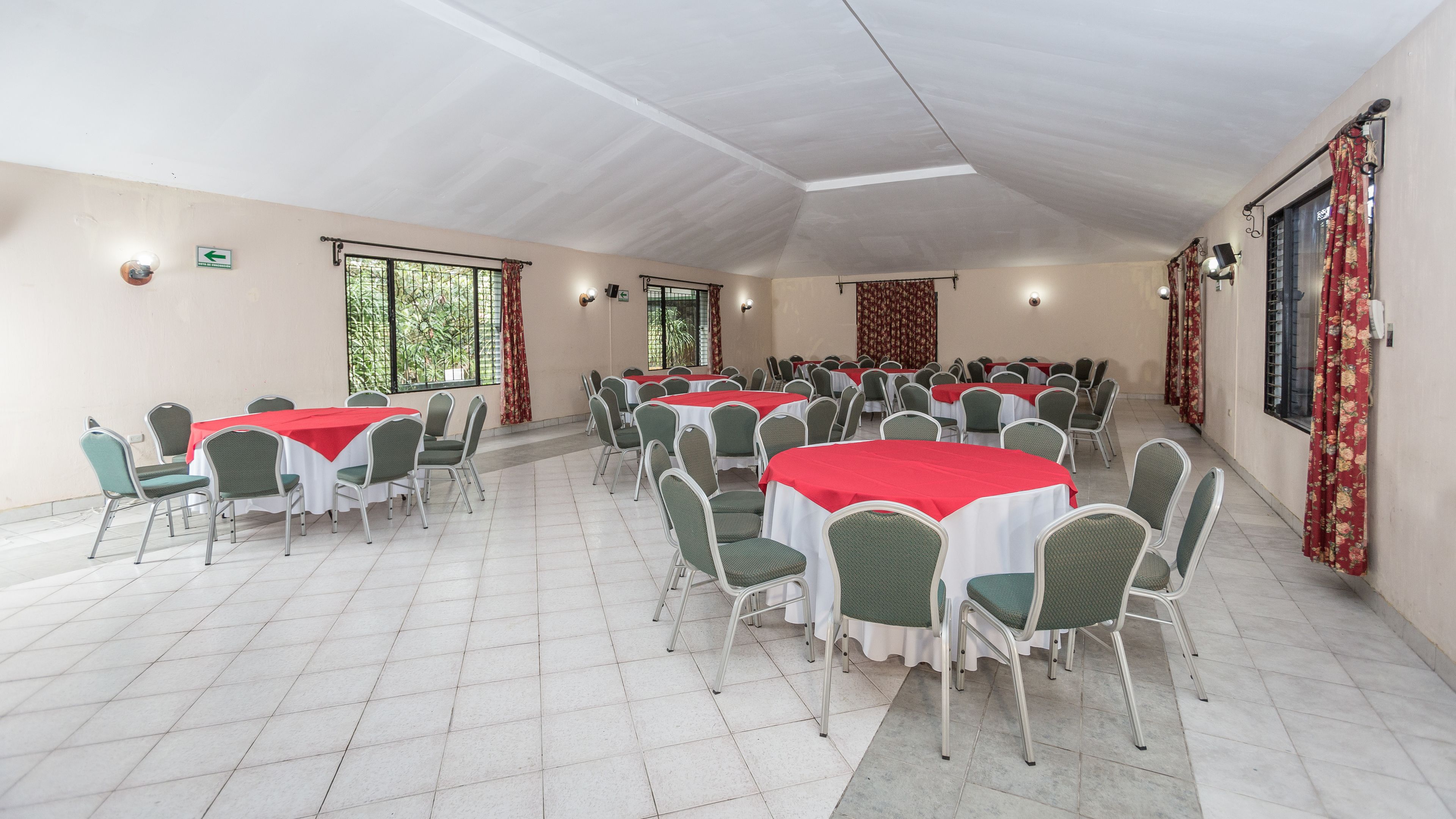 Un salón amplio con varias mesas redondas cubiertas con manteles rojos y sillas verdes.