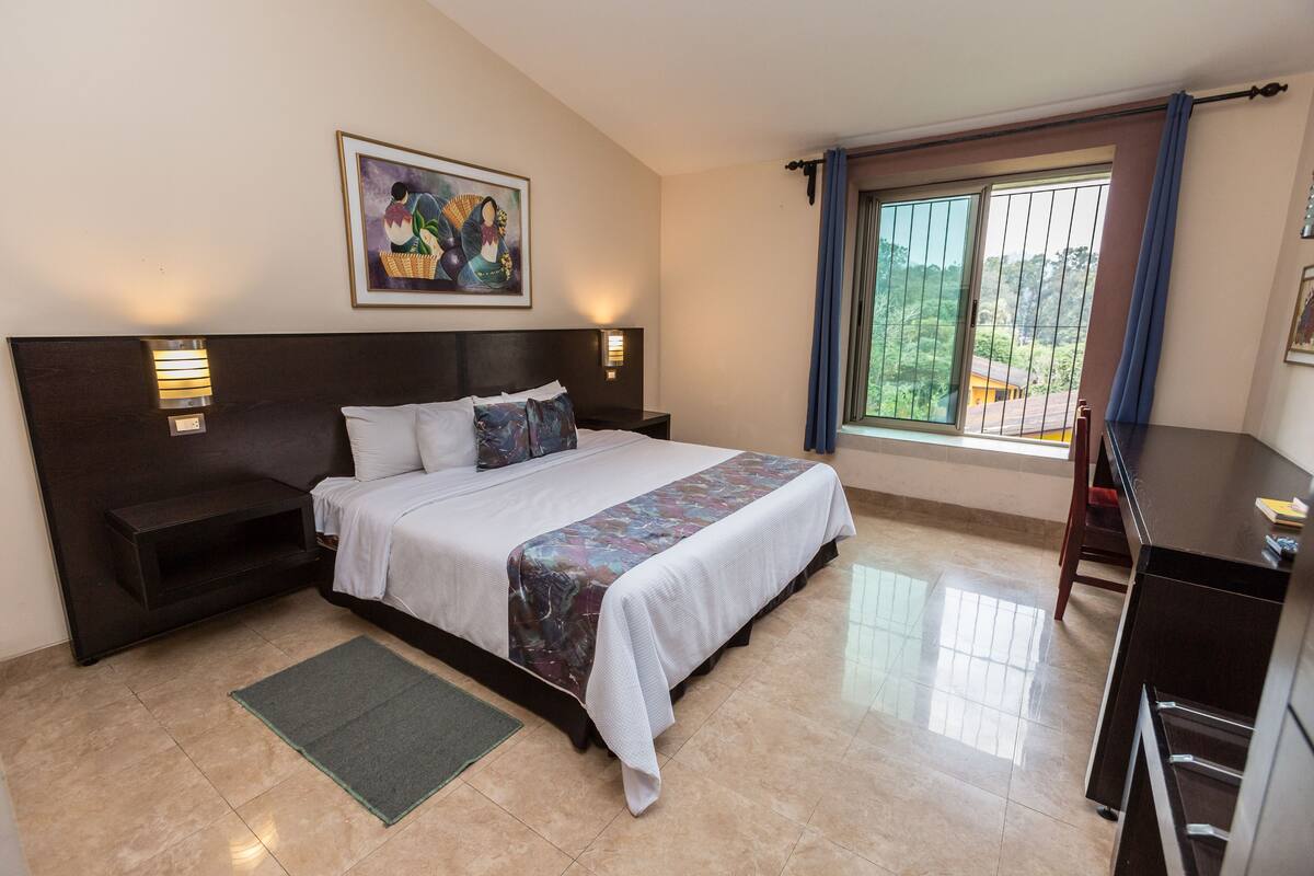 A neatly arranged bedroom with a large bed, a desk, and a window with a view of greenery.