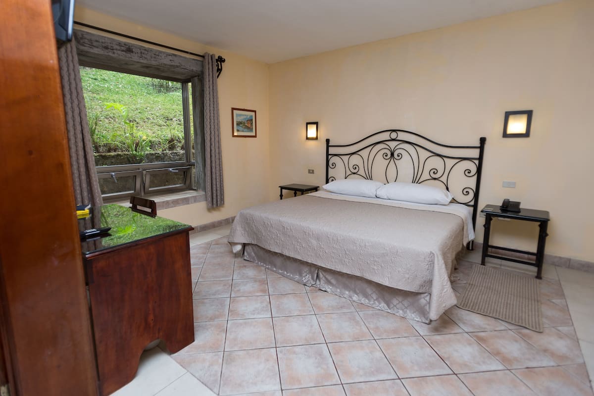 A bedroom with a bed, two bedside tables, a nightstand, a wooden desk, a window with curtains, and a small picture on the wall.
