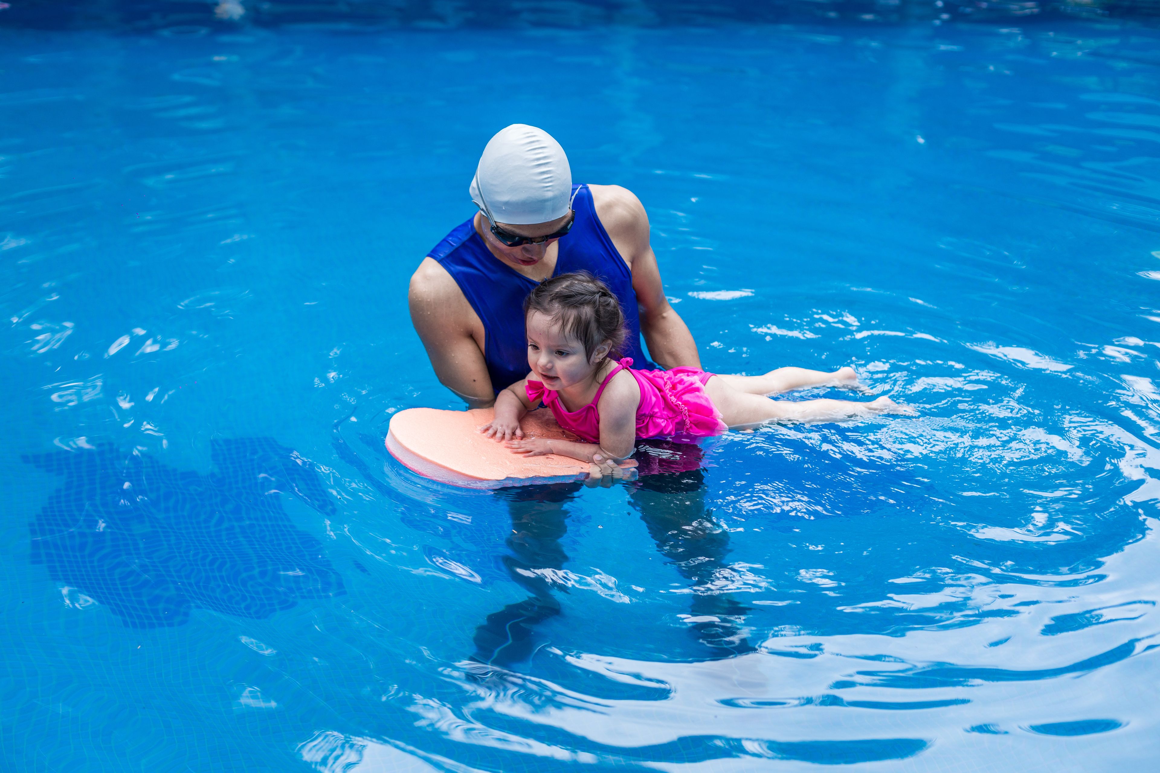 A person in a blue swimsuit and swim cap assisting a child in a pink swimsuit on a pool float.