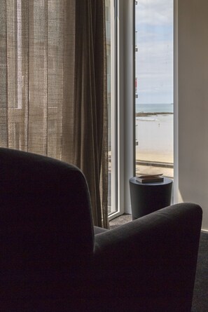 A room with a view of the beach through a window, a chair, and a small table with a book. - Hôtel L'Adresse (Saint-Malo)