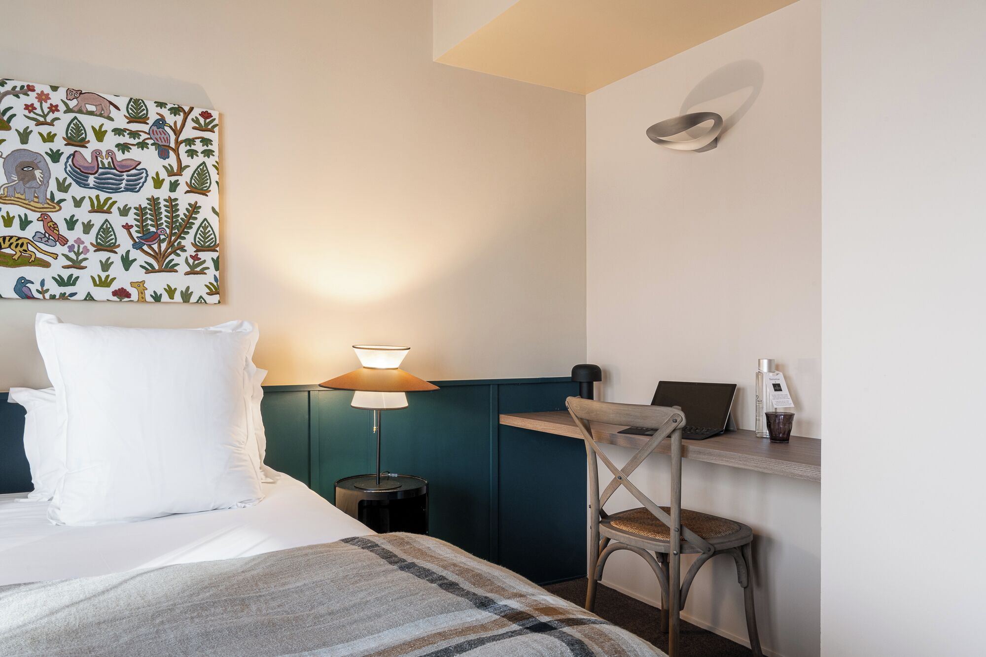 A hotel room with a bed, a desk, a chair, a lamp, and a wall with a colorful mural.
