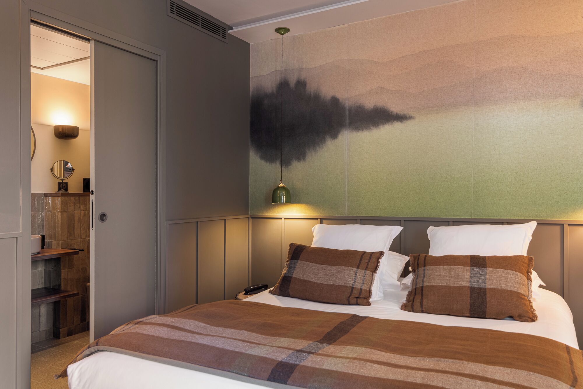 A hotel room with a bed, a nightstand, a wall-mounted lamp, and a view of the outdoors.