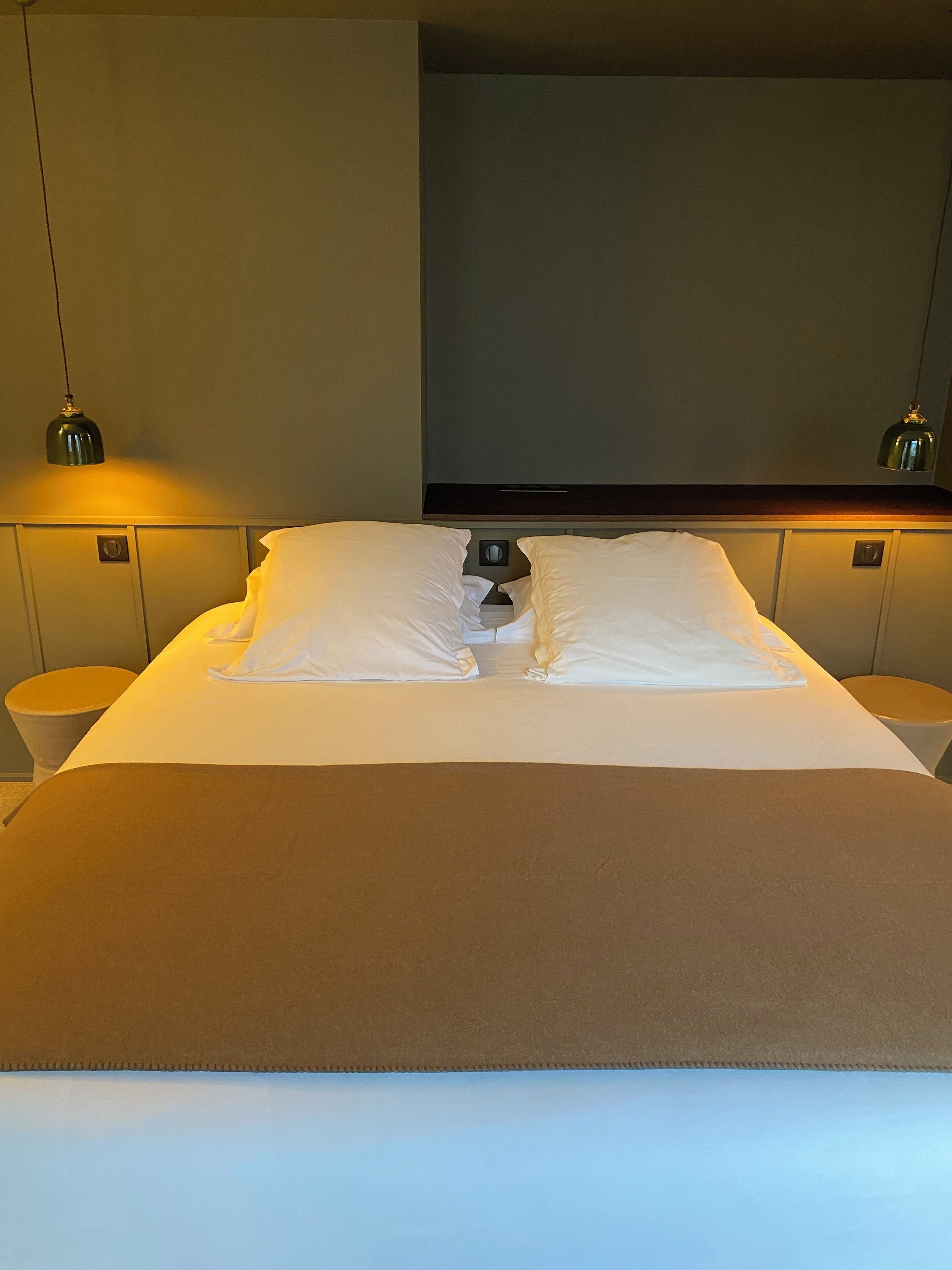 A neatly made bed with white linens and a brown accent, two bedside lamps, and a wall-mounted TV.
