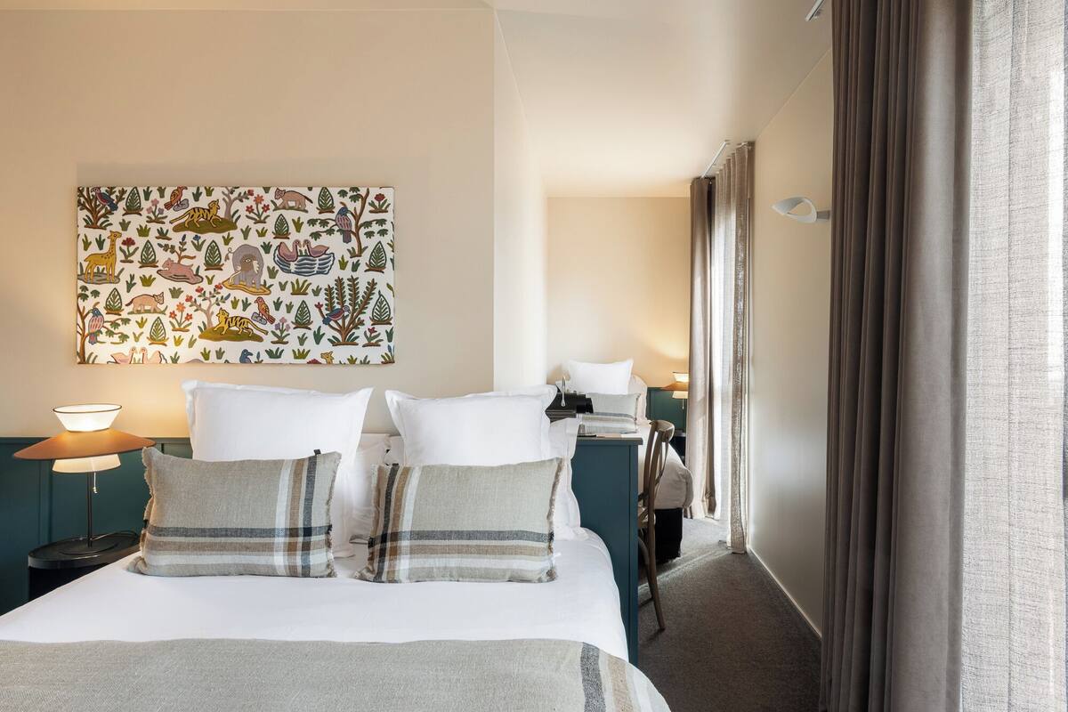 Ein Hotelzimmer mit Bett, Nachttisch, Lampe, einem Bild an der Wand und einem Fenster mit Vorhängen.