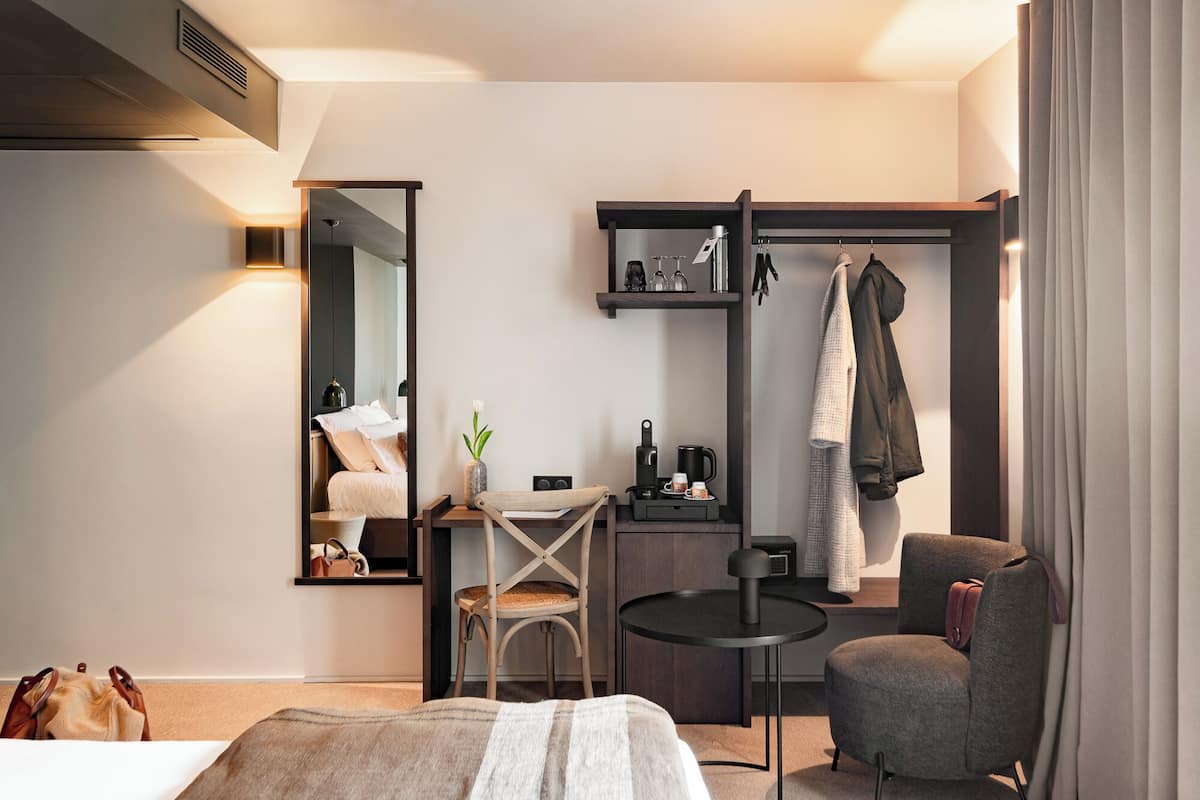 Ein modernes Hotelzimmer mit Bett, Schreibtisch mit Stuhl, einem kleinen runden Tisch, einem Spiegel, einem Kleiderschrank und einer Sitzecke mit Sessel.