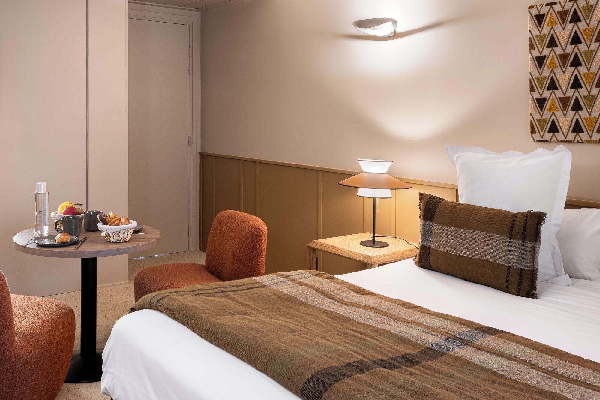 A hotel room with a bed, a nightstand, a lamp, a chair, a small table with breakfast items, and a wall with a geometric pattern.