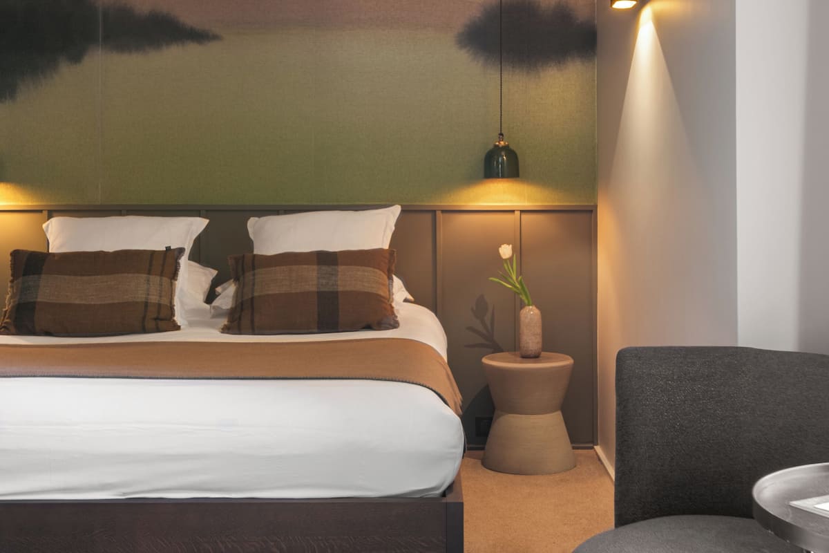 Ein modernes Hotelzimmer mit einem großen Bett, einem kleinen runden Tisch, einem grauen Sessel und einer Wandleuchte.