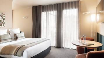 A hotel room with a bed, a round table with a book and glasses, a chair, and a large window with curtains.