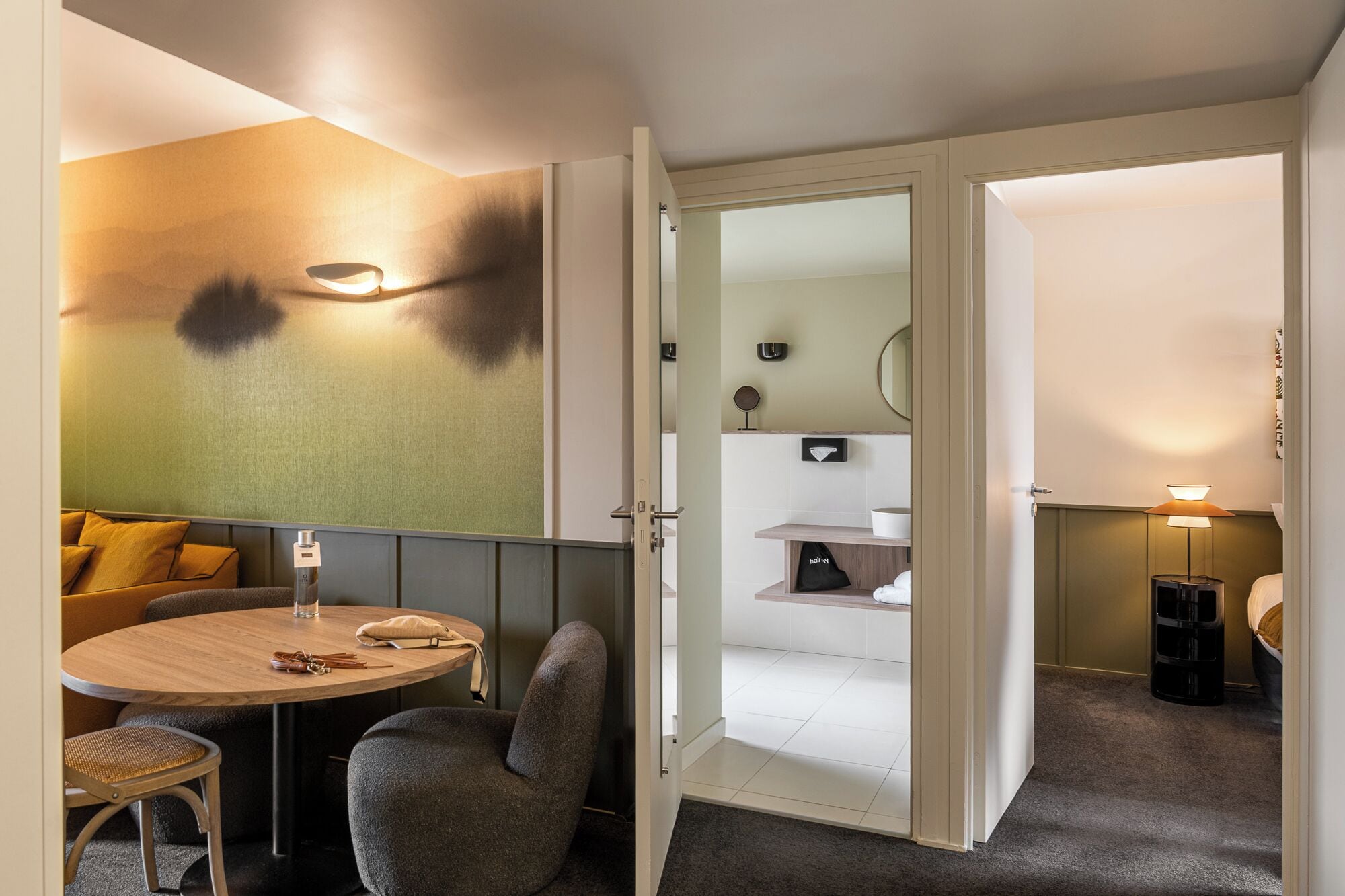 A modern hotel room with a round wooden table, a grey armchair, a sofa with yellow cushions, a small round mirror, and a bedside lamp.