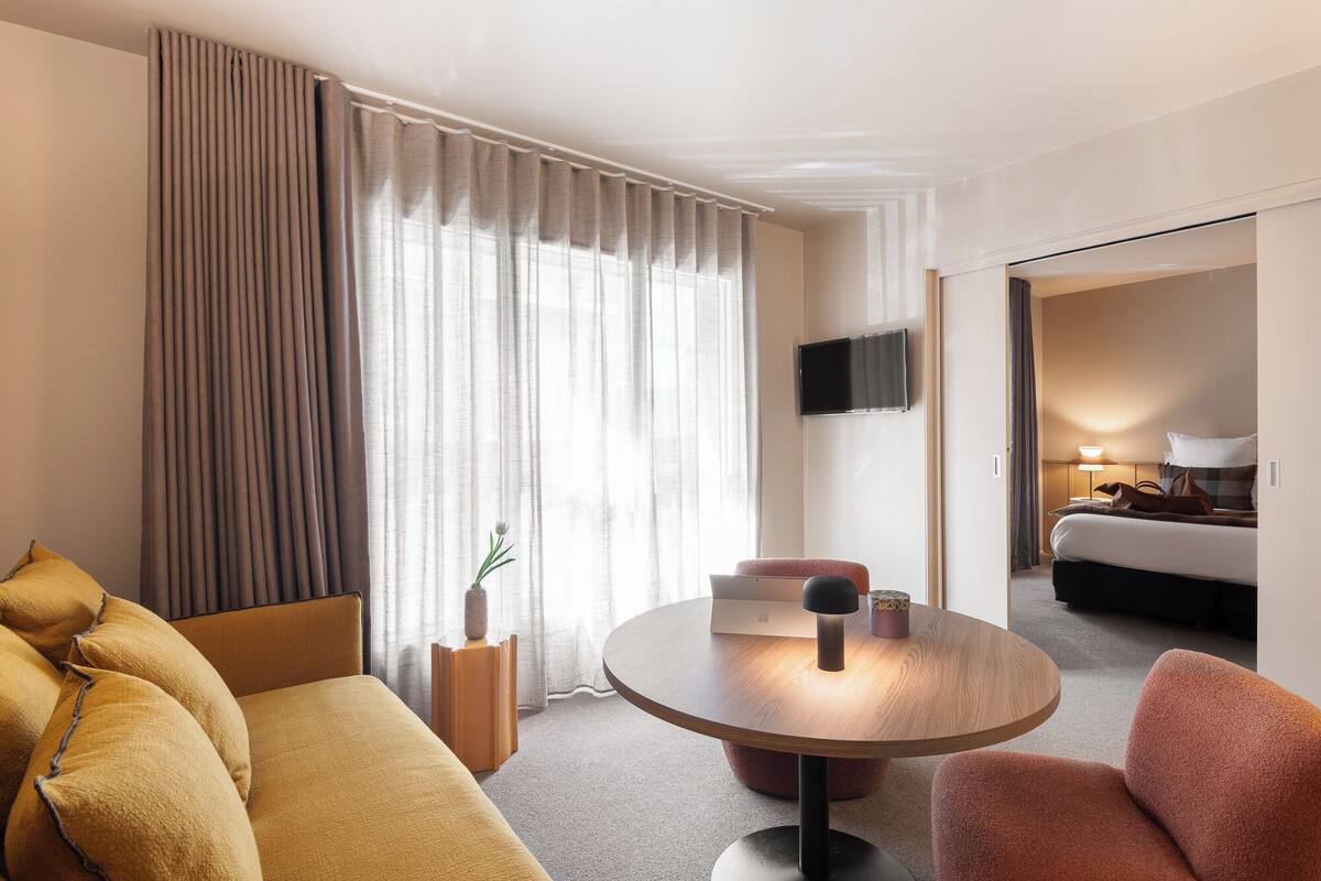 Ein Hotelzimmer mit einem Bett, einem runden Tisch mit Lampe, einem an der Wand befestigten Fernseher und einer Couch mit Kissen.