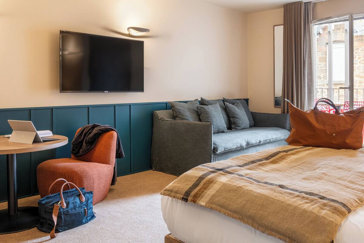 Ein Hotelzimmer mit Bett, einer Couch, einem Schreibtisch mit Laptop, einem Fernseher und einem Fenster mit Vorhängen.