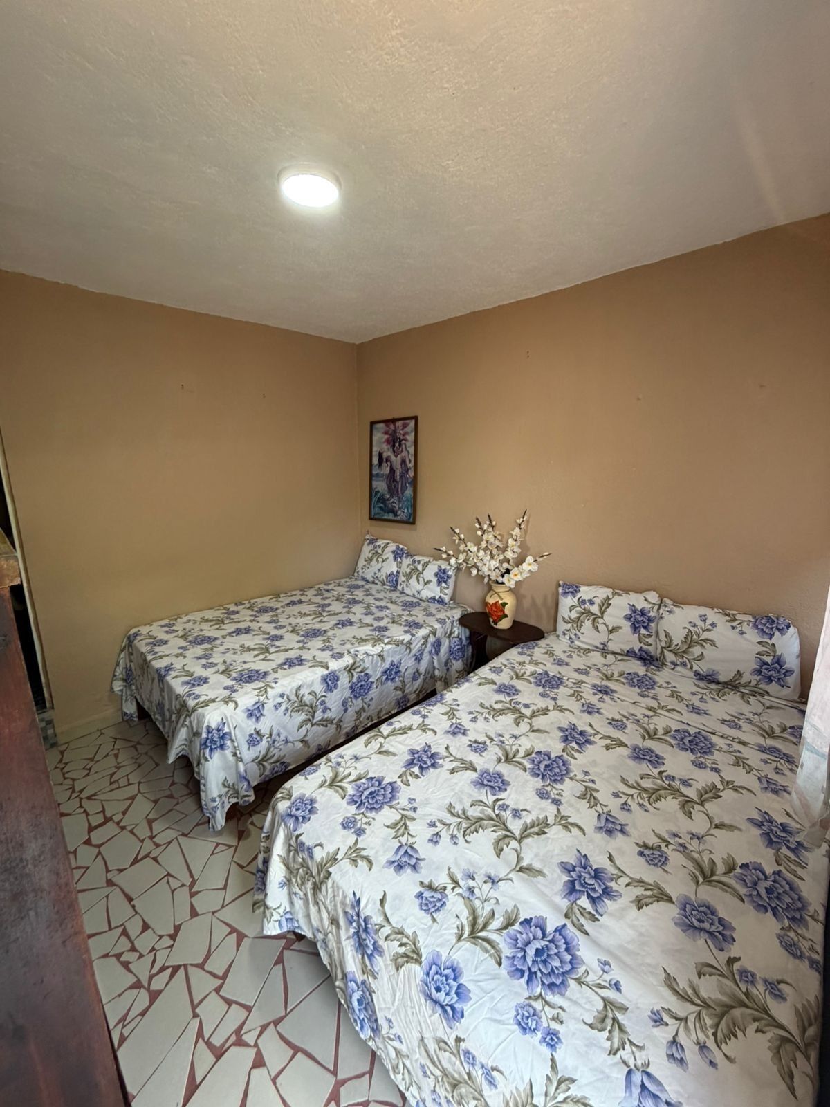 A bedroom with two beds, a small table with a vase, and a painting on the wall.