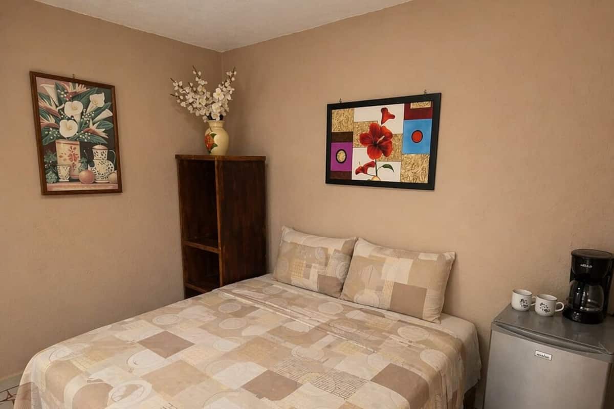 A bedroom with a bed, a small refrigerator, a coffee maker, and two wall-mounted paintings.