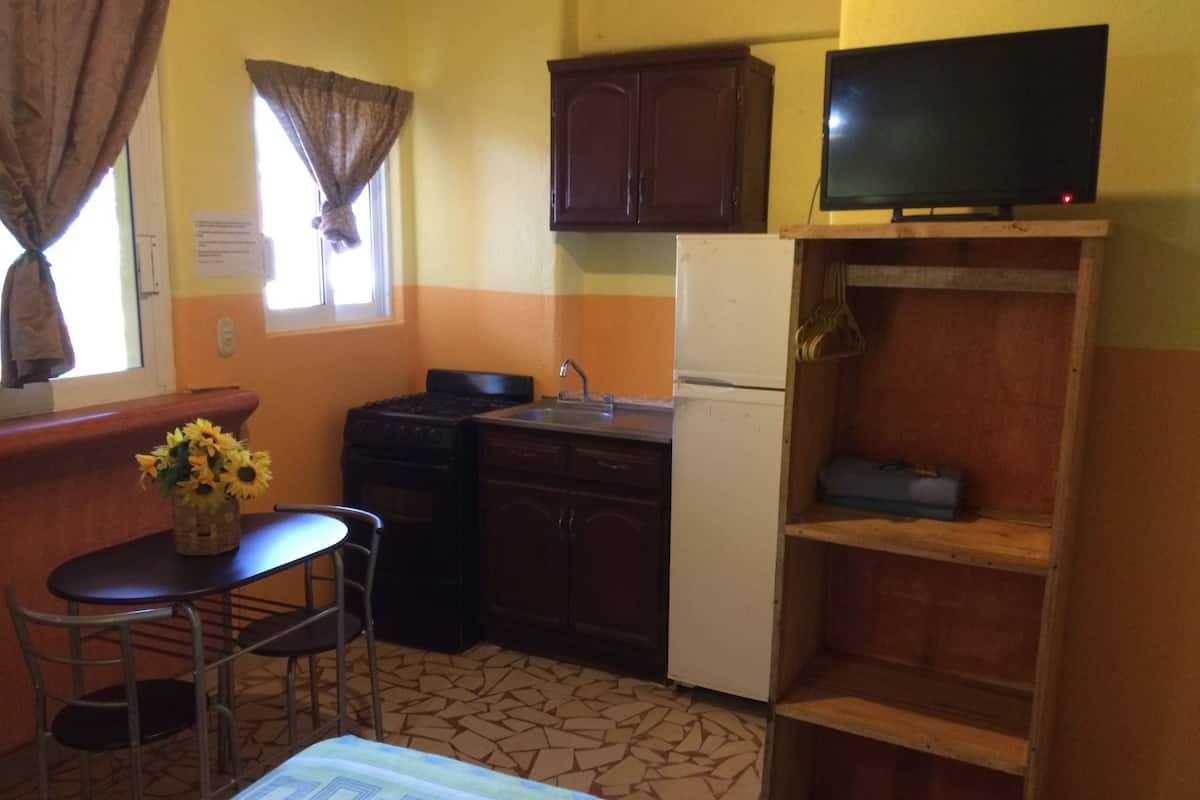 A compact kitchen with a stove, sink, and refrigerator, a small dining table with chairs, and a television on a shelf.