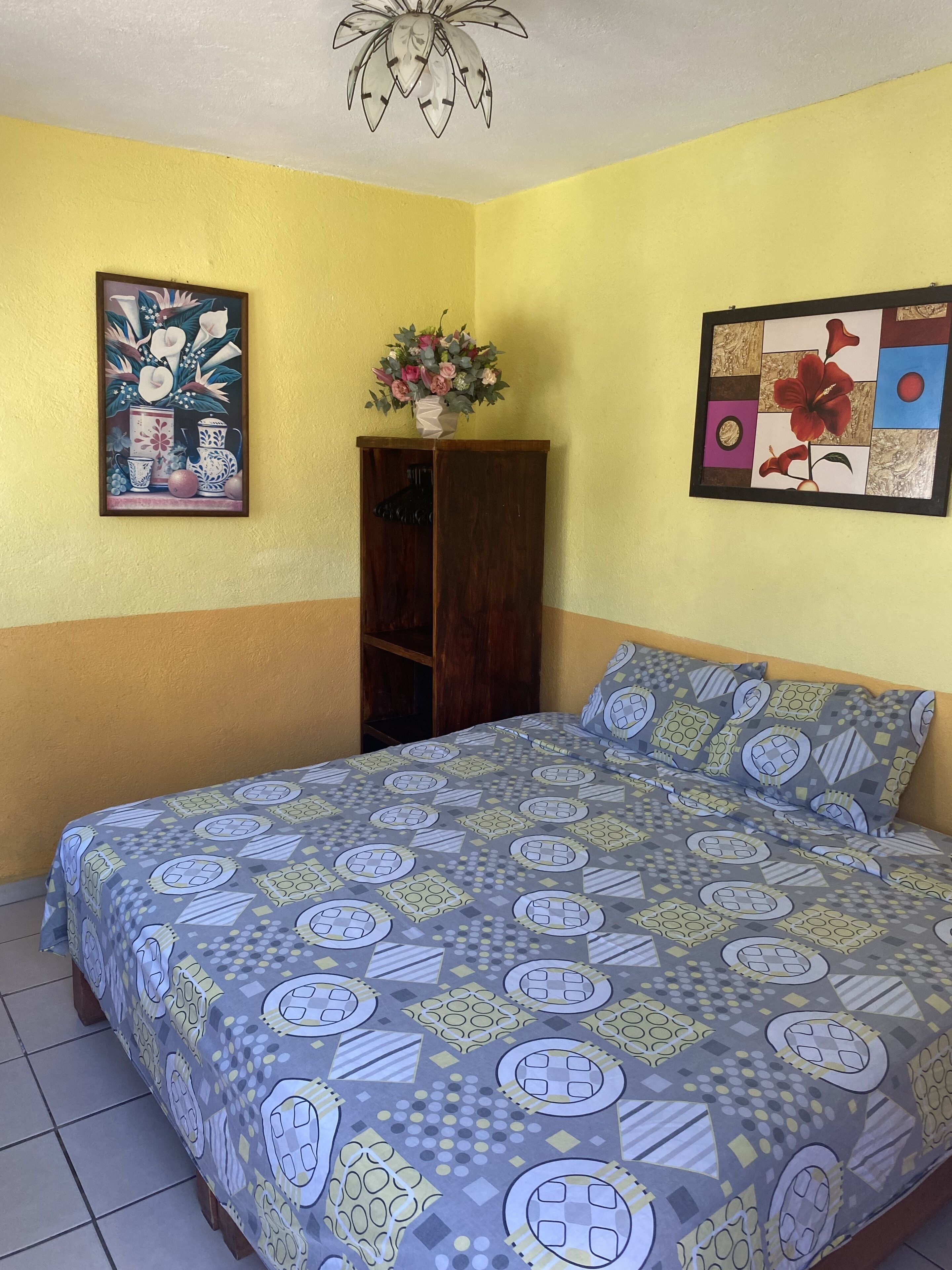 A bedroom with a bed, a wooden wardrobe, a vase with flowers, and two framed artworks on the walls.