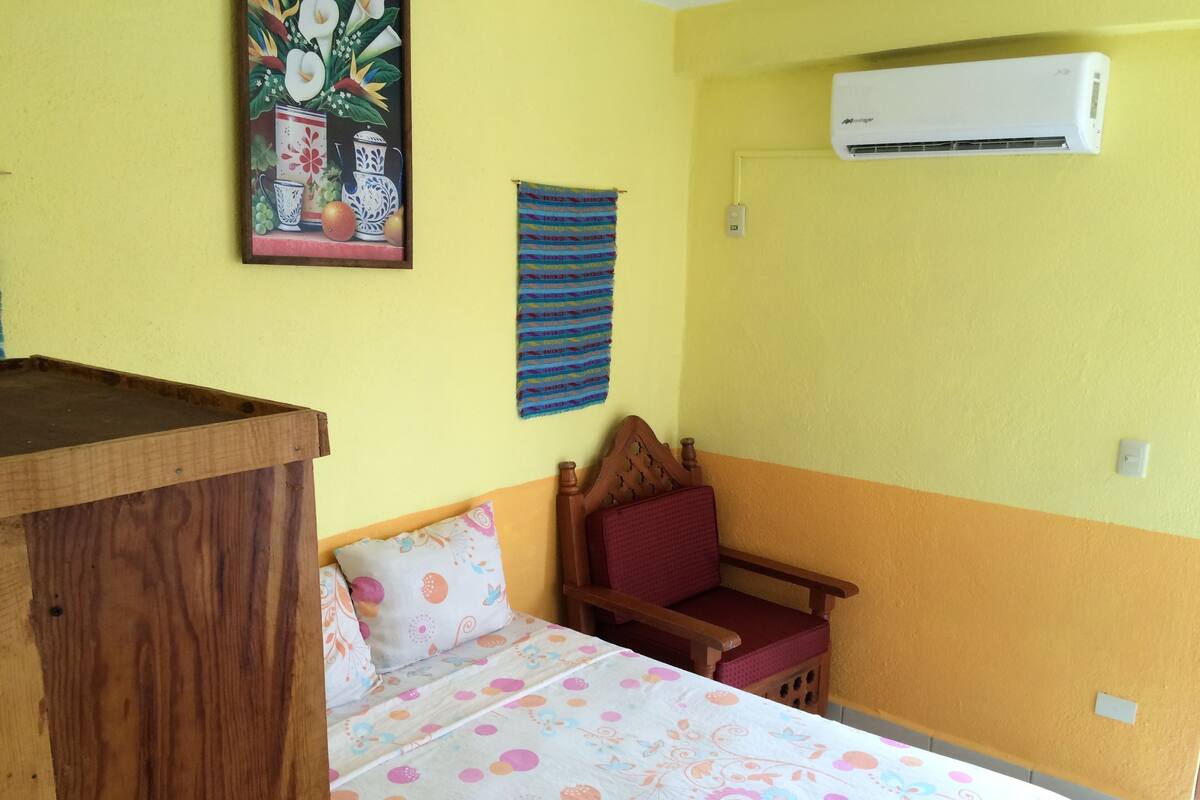 A room with a bed, a wooden desk, a chair, a wall-mounted air conditioner, and a colorful painting.