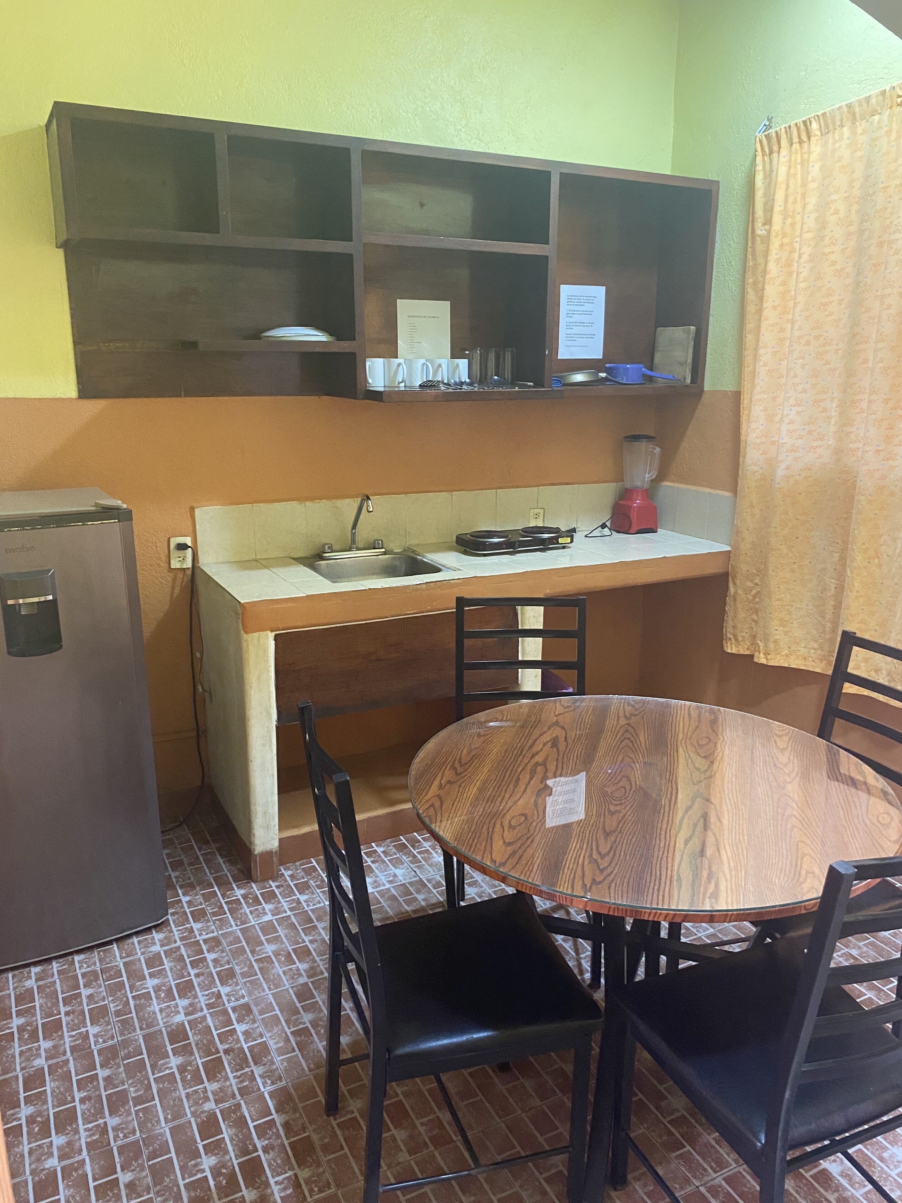 A compact kitchen with a refrigerator, a sink, a stove, and a blender on the counter. There is a round wooden table with chairs and a shelf above the counter.