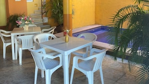 Un patio al aire libre con sillas y mesas de plástico blanco, una pared amarilla, techo de madera y un área de piscina.