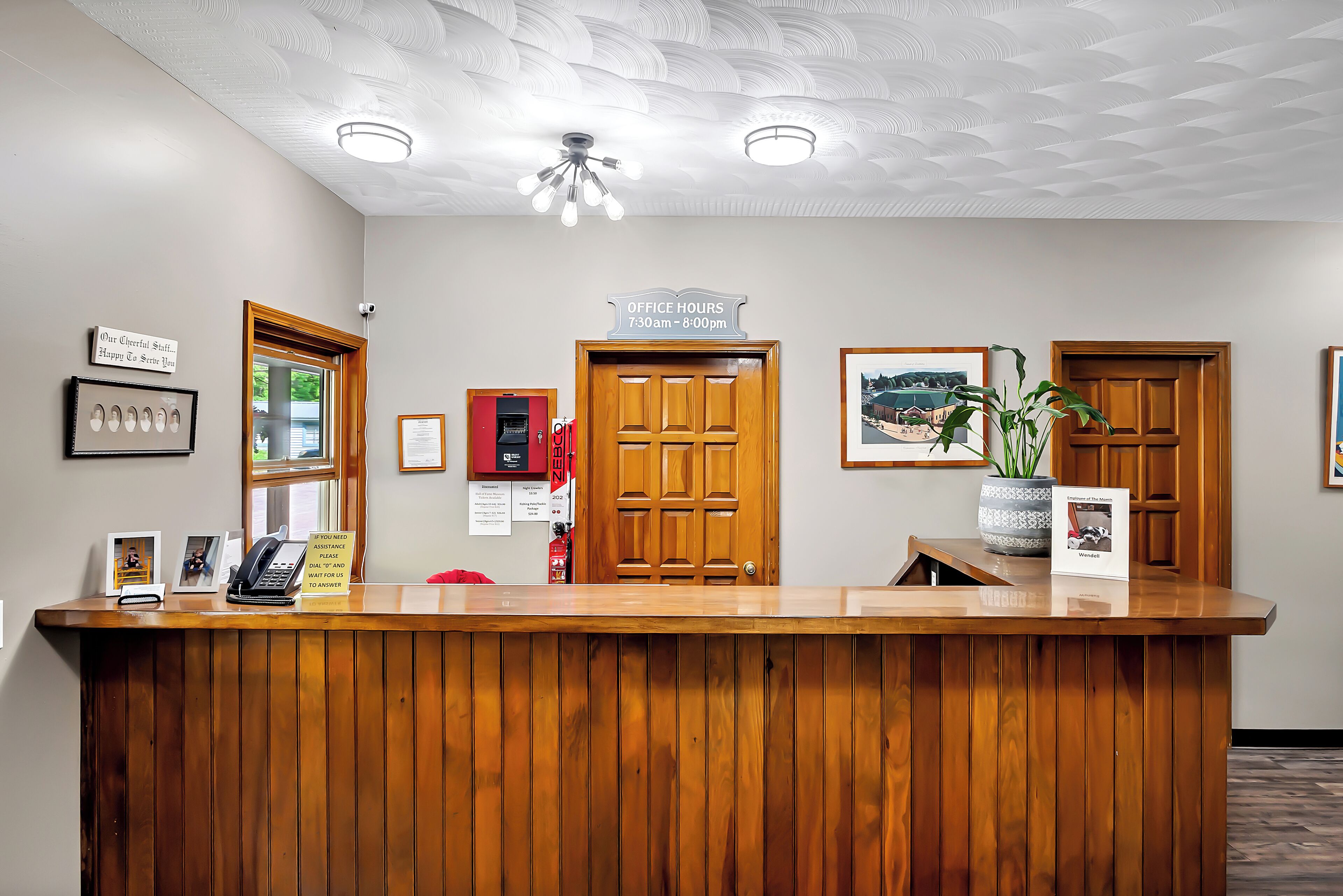 A reception desk with a wooden finish, a sign indicating office hours, framed pictures, a potted plant, and a fire extinguisher.