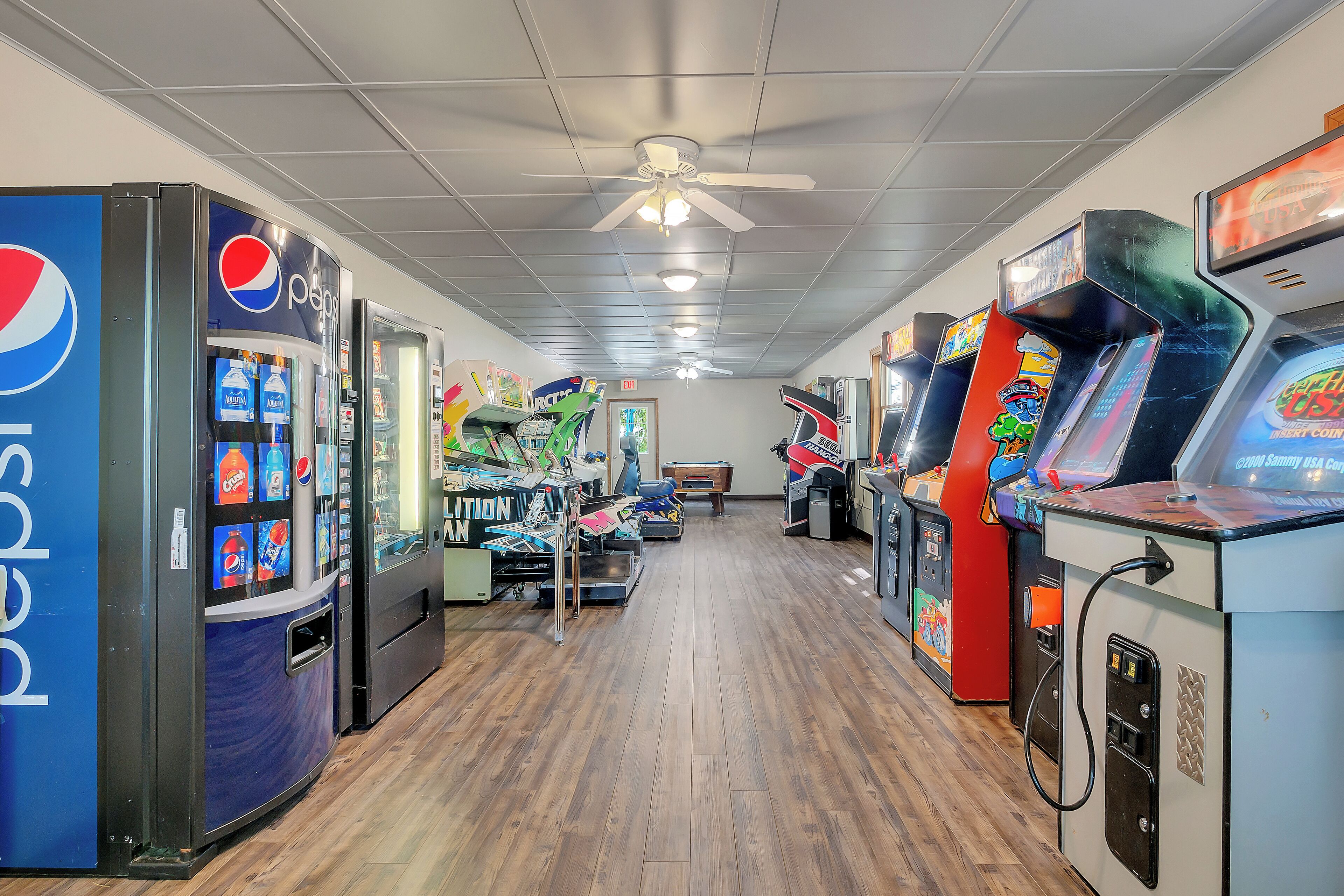Une rangée de jeux d’arcade et un réfrigérateur Pepsi dans une pièce au plancher en bois et dotée d’un ventilateur de plafond.