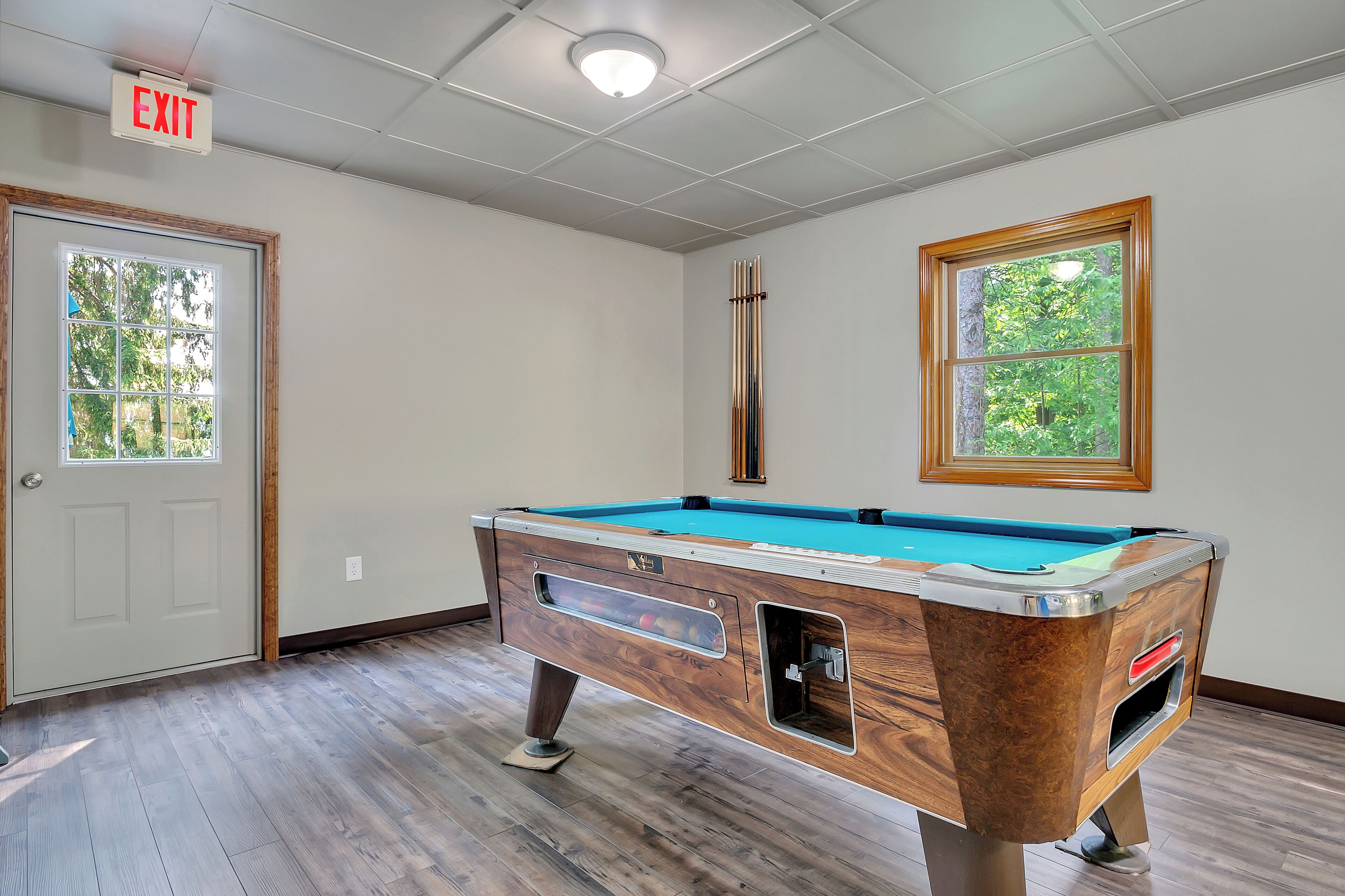 A pool table in a room with wooden flooring, a door, and a window with a view of trees.